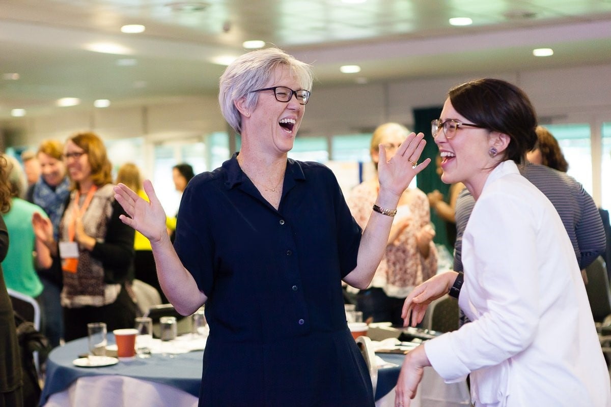 Two workshop participants laughing together while standing among other attendees.